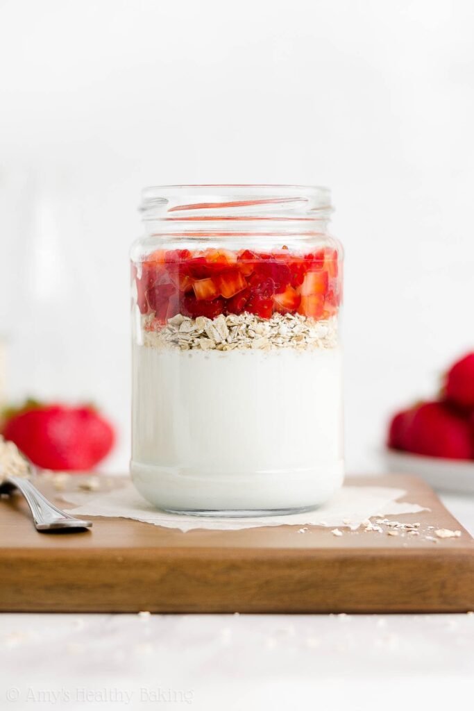 a glass jar holding the layered ingredients used to make healthy strawberry protein overnight oats, while resting on a wooden cutting board alongside a spoonful of oats – easy overnight oatmeal recipe – high protein, gluten free, sugar free, low fat, clean eating – made with Greek yogurt, oats, fresh strawberries, and no added sugar